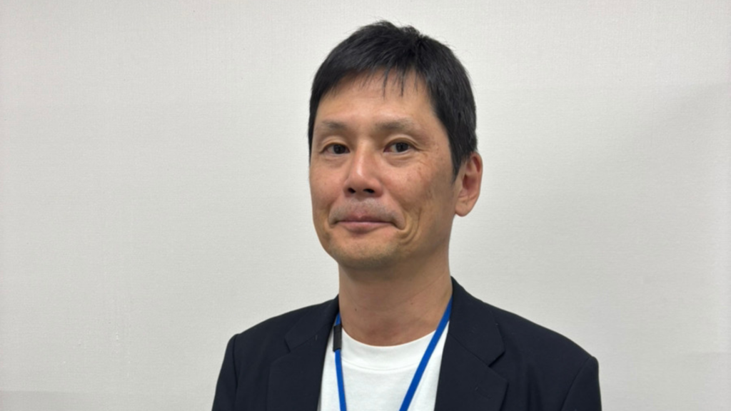 アイ・ワークス代表、田村の顔写真。黒のジャケットに白のTシャツ、首から社員証をかけ、穏やかな表情で正面を向いているポートレート。