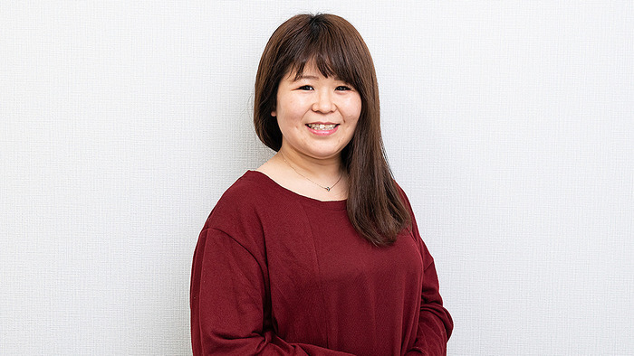 アイ・ワークススタッフ、青柳の写真。茶色のロングヘアで、エンジ色のトップスとシルバーのネックレスを着用し、明るい笑顔で正面を向いているポートレート。