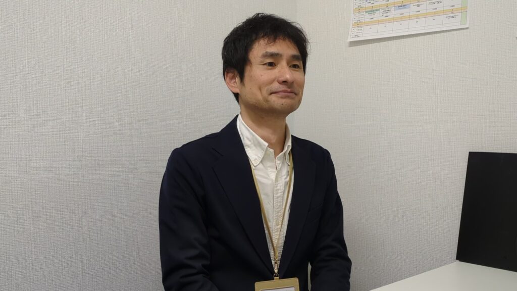 アイ・ワークススタッフ、小池の写真。濃紺のジャケットと白シャツを着用し、首から社員証を下げ、穏やかな表情で少し右前を見ている様子。