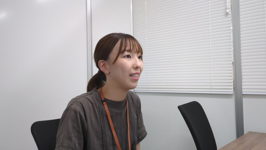 アイ・ワークス サービス管理責任者、竹中の写真。髪を後ろでまとめ、ダークグレーのトップスとオレンジ色のネックストラップを着用し、ブラインドのあるオフィスで笑顔で話している様子。