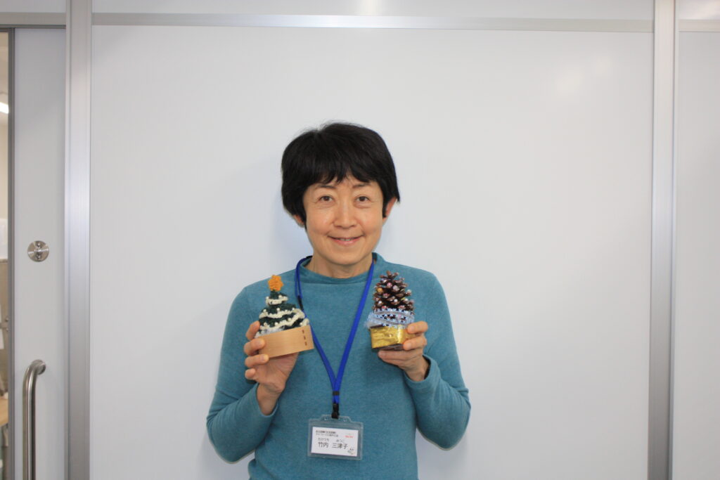 アイ・ワークススタッフ、竹内の写真。ショートヘアで青緑色のトップスを着用し、両手に松ぼっくりで作った手作りの飾りを持って、優しく微笑んでいる正面のポートレート。